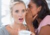 Two women in a casual setting, one whispering to the other who looks surprised
