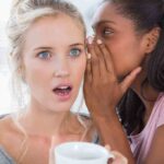 Two women in a casual setting, one whispering to the other who looks surprised