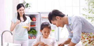 A father and daughter happily preparing vegetables in a bright kitchen, while a woman observes.