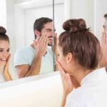 A couple smiling at their reflections in a bathroom mirror while applying skincare products