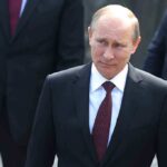 Putin Parades NEW NUKE – Military Experts Worried A serious-looking man in a suit walking outdoors with security personnel in the background
