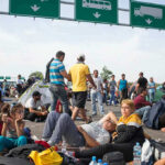Refugees Axed First—Americans IMPACTED Next! People sitting and resting on a highway.