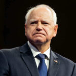 Walz Pull Race Card Blame In Shocking Fraud Scandal Defense Man in suit with a U.S. flag pin.