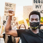 Black Panthers Return – Armed Resistance RISING Protester raises fist, wears mask, signs in background.