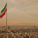 Iran MASSACRES 544 Protesters – BLOODBATH Unfolds Iranian flag waving over a city skyline with mountains in the background