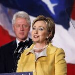 Clintons SUBPOENAED – Ordered to Testify Hillary Clinton delivering a speech with Bill Clinton in the background