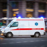 Paramedics On The Hook For Brutal Murder Ambulance driving on urban street with lights flashing.