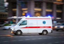 Paramedics On The Hook For Brutal Murder Ambulance driving on urban street with lights flashing.