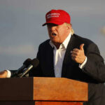 Trump Backed Candidate FALLS In Primary Shocker! Person speaking at a podium wearing a red hat.