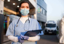 Nurse FIRED – Vowed To Not Treat Conservatives! Nurse in scrubs and mask outside hospital holding clipboard.