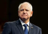 Tim Walz Pardons WORST of the WORST Illegal! Man in suit with a U.S. flag pin.