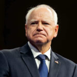 Tim Walz Pardons WORST of the WORST Illegal! Man in suit with a U.S. flag pin.