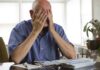 Elderly man sitting at a table with his hands covering his face, surrounded by paperwork