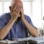 Elderly man sitting at a table with his hands covering his face, surrounded by paperwork