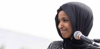 Ilhan Omar speaking at a microphone with hand on chest