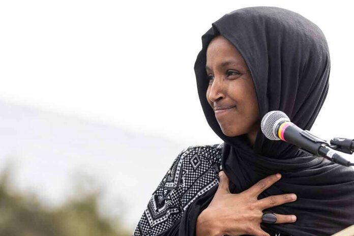 Ilhan Omar speaking at a microphone with hand on chest