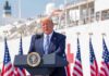Donald Trump speaking at a podium with American flags and a ship in the background