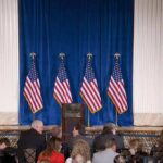 Dem Candidate Turns Back on Flag – Outrage Sparks! Empty podium on stage with multiple U.S. flags and audience waiting