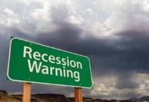 Green roadside sign reading 'Recession Warning' under stormy sky