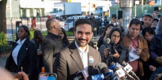 Zohran Mamdani speaks at a press conference; Julia Bacha is visible among the crowd