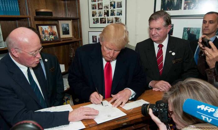 Donald Trump signs a document as Corey Lewandowski looks on amid officials and press
